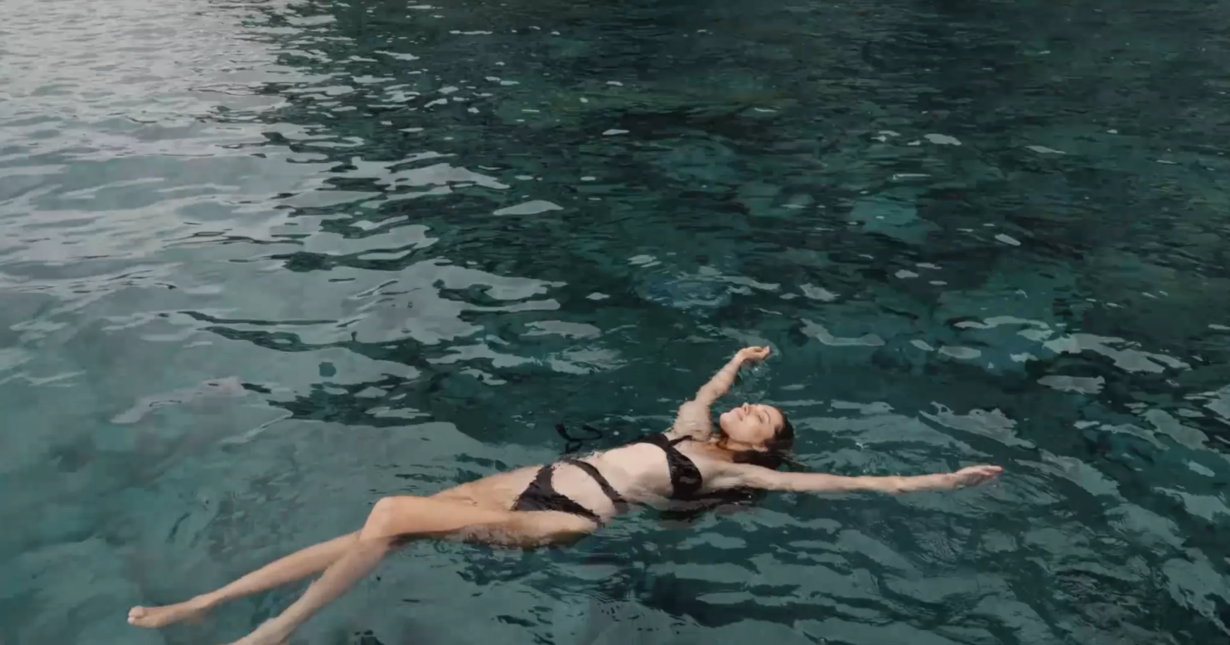 Una donna in bikini nero galleggia a pancia in su in un'acqua limpida e calma, con le braccia e le gambe rilassate.