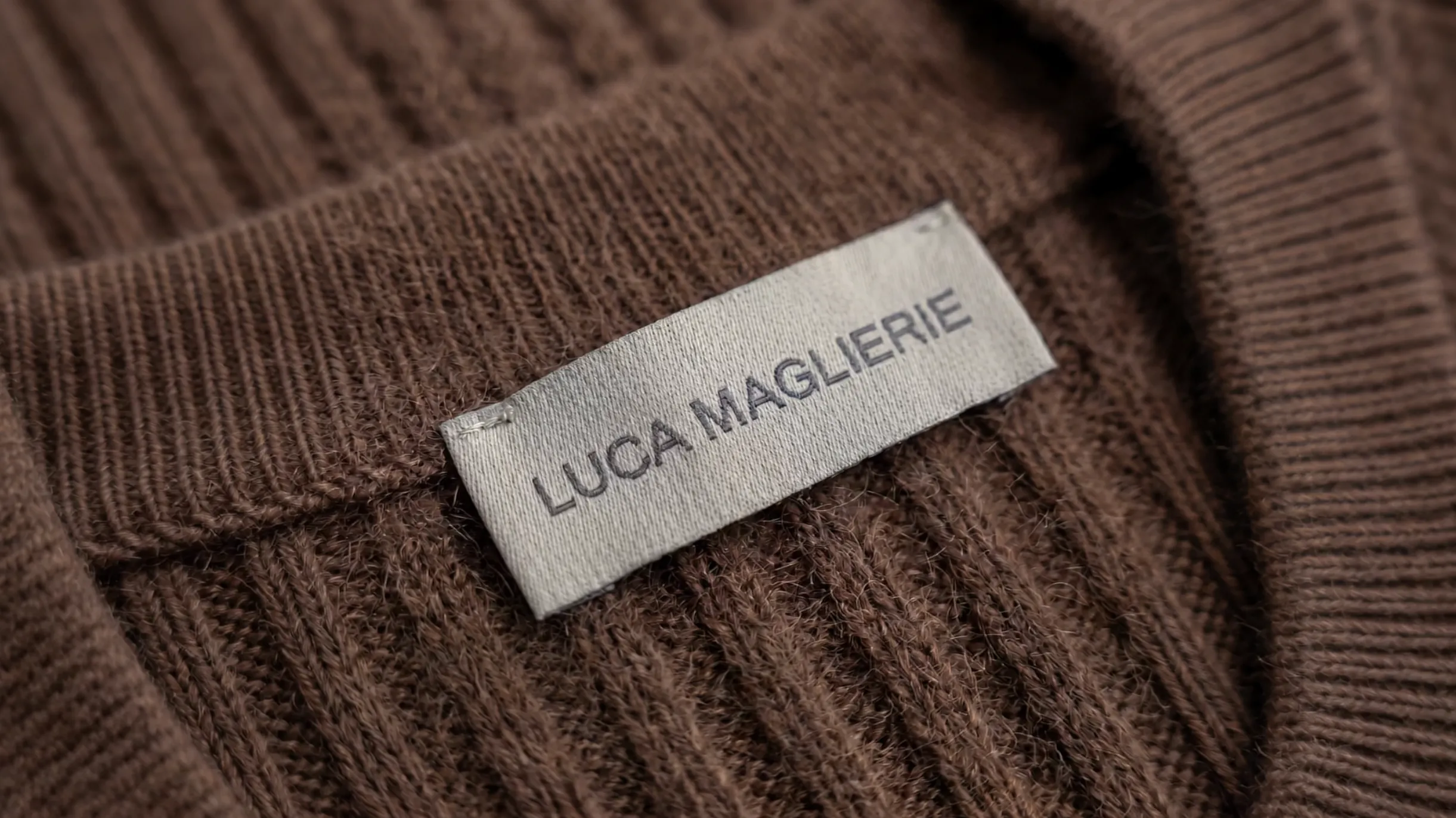 Primo piano di un'etichetta bianca cucita su un capo in maglia marrone con trama a coste, che riporta la scritta 'LUCA MAGLIERIE' in lettere maiuscole.